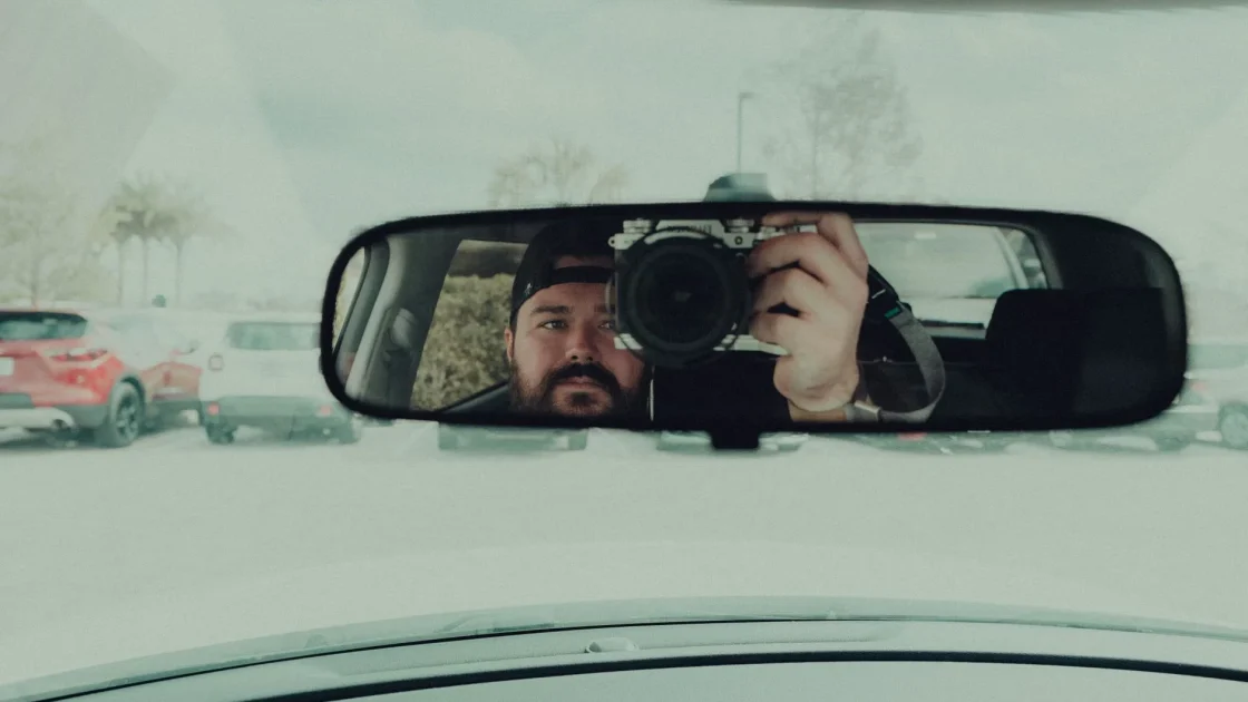 A pedestrian crossing a street ahead of a car, viewed through the windshield with the forward ADAS camera housing visible near the mirror.