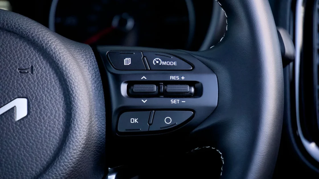 Close-up of a steering wheel showing the cruise control buttons with a small car and distance-bar icon on the dashboard behind it.