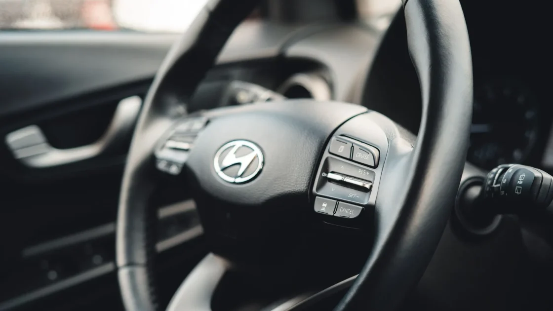 A driver's hand near the adaptive cruise control buttons on a steering wheel with the road ahead in view.