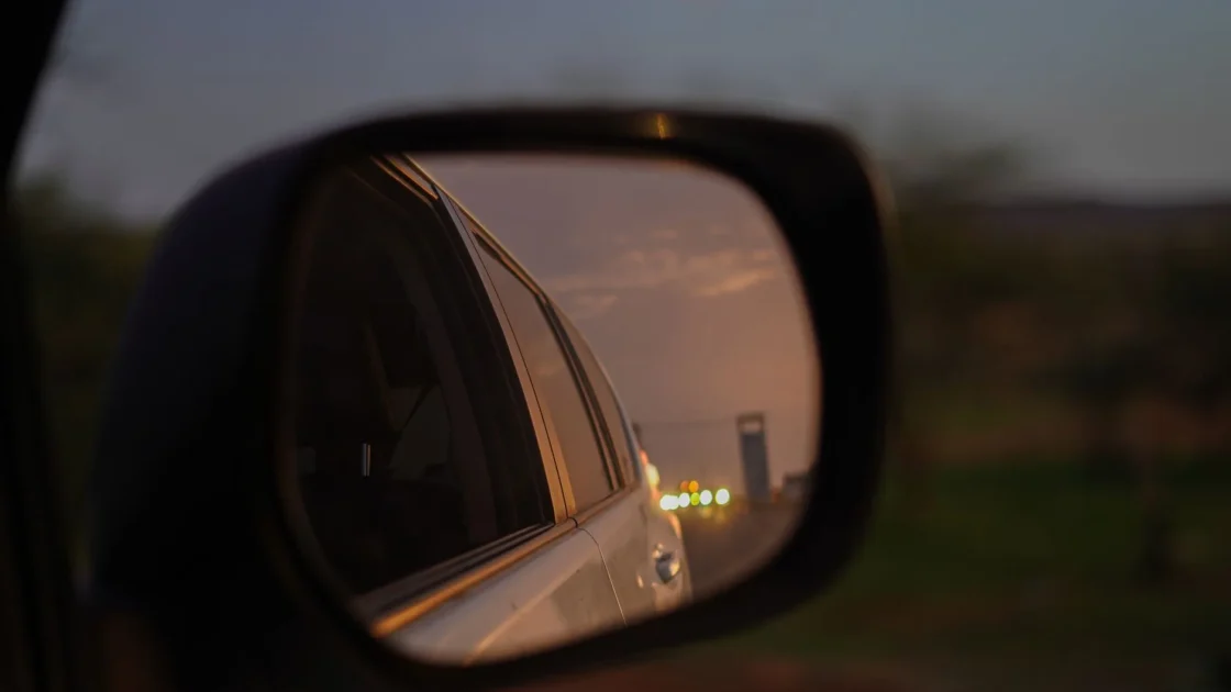 Close-up of a car side mirror with an amber blind spot warning symbol lit as another vehicle sits in the adjacent lane.