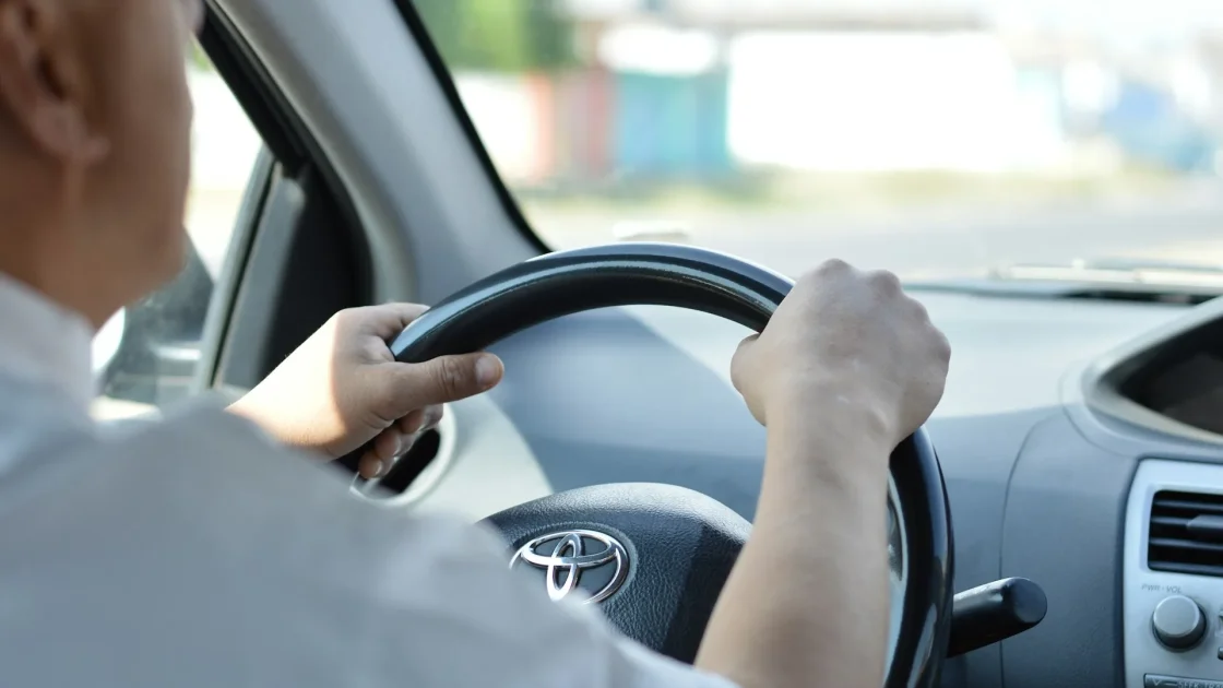 A driver's hands on the wheel of a car with the lane keep assist indicator lit on the instrument cluster.