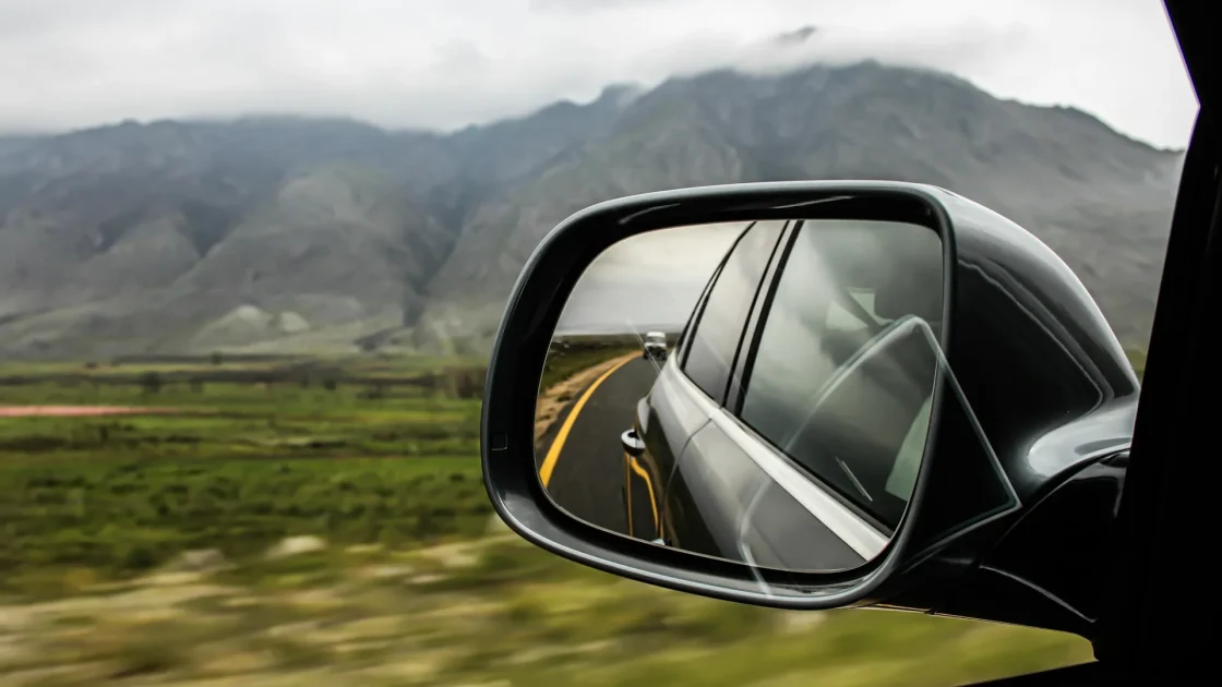 A car side mirror with an illuminated amber blind spot warning icon in the upper corner of the glass.