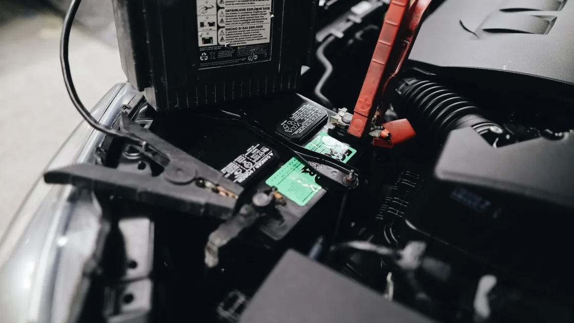 Jumper cables clamped to a car battery terminal with the hood open during a jump start.